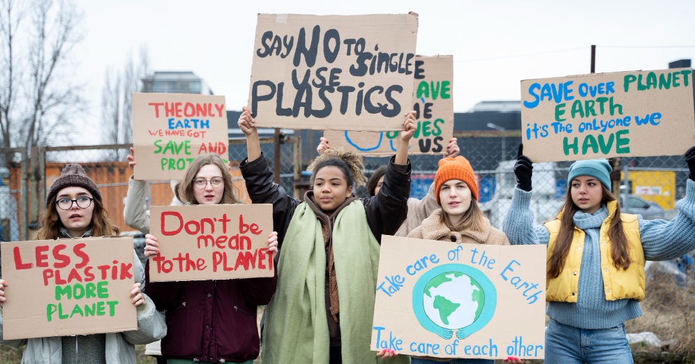 A picture of women in front of a chain link fence holding up cardboard signs calling for people to take care of the planet and reduce plastic use. 